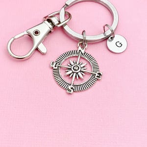 May include: Silver compass keychain with a silver split ring and a silver lobster clasp. The compass has a silver star in the center and the letters N, S, E, and W around the edge. The keychain also has a small silver charm with the letter G on it.