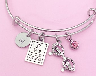May include: Silver charm bracelet with a letter M charm, eye chart charm, and glasses charm. The bracelet also has a small pink rhinestone charm.