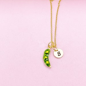 Peut inclure: Un collier dor&eacute; avec un pendentif en forme de gousse de pois verte et une breloque ronde avec la lettre "S". Le pendentif contient des pois jaunes. Le collier est pr&eacute;sent&eacute; sur un fond rose.