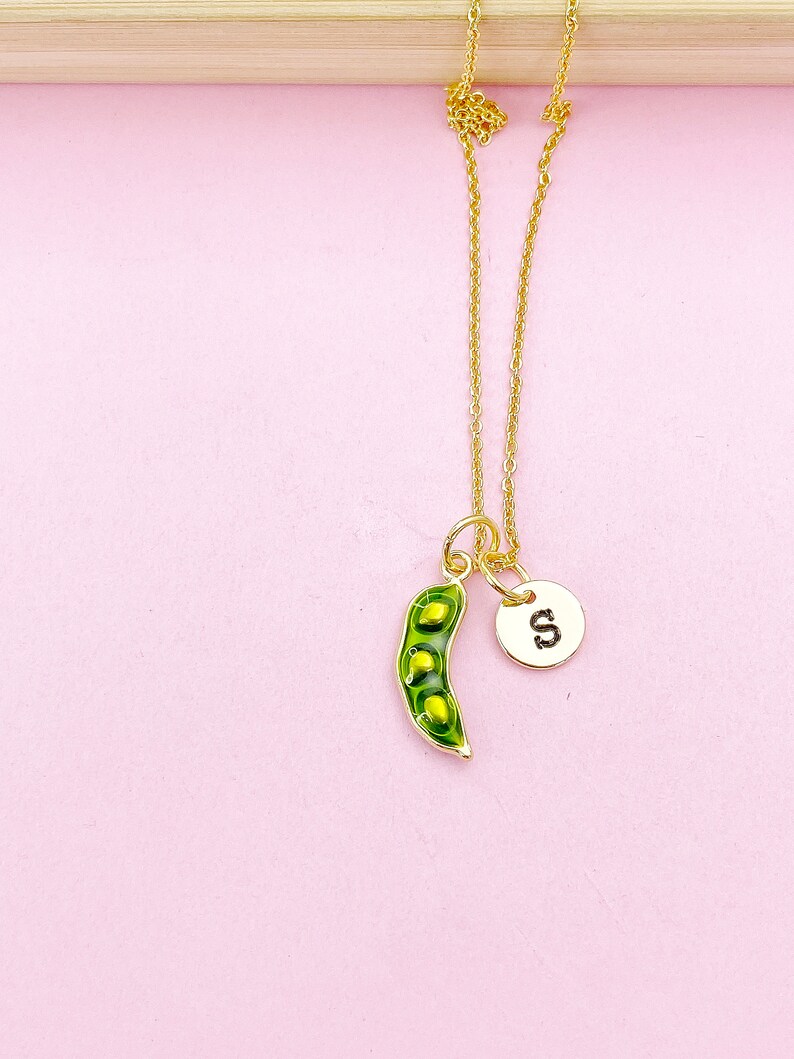 Peut inclure: Un collier dor&eacute; avec un pendentif en forme de gousse de pois verte et une breloque ronde avec la lettre "S". Le pendentif contient des pois jaunes. Le collier est pr&eacute;sent&eacute; sur un fond rose.