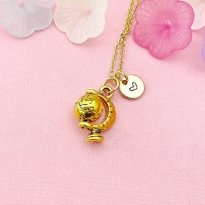 May include: A gold-colored necklace with a globe pendant and a small charm with a heart symbol. The necklace is on a pink surface, surrounded by colorful flower-shaped decorations.