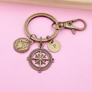 May include: A gold-toned keychain with a compass, a globe, and a small charm with the letter "P" engraved on it. The keychain is on a pink background.