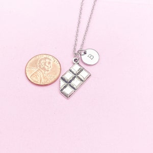 May include: A silver necklace with a chocolate bar charm and a silver charm with the letter "B" on it. The necklace is on a pink background.