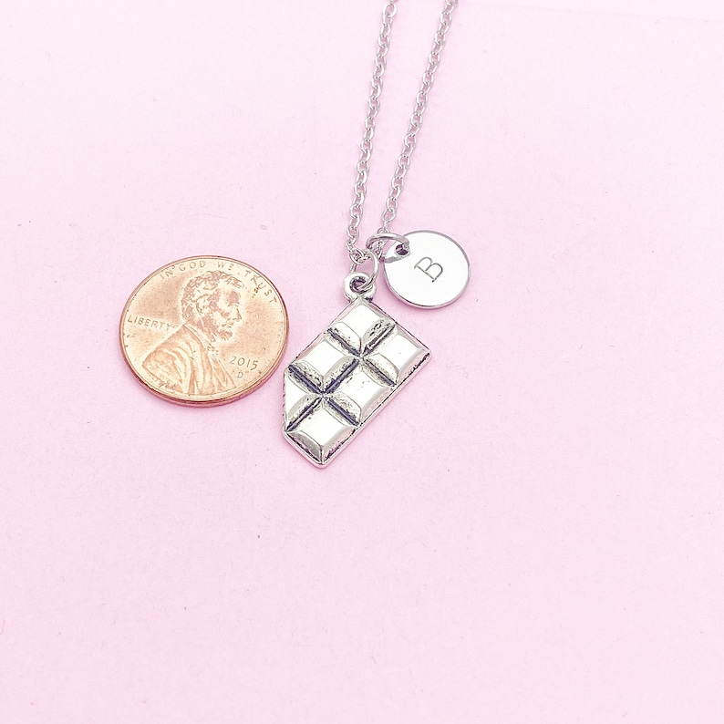 May include: A silver necklace with a chocolate bar charm and a silver charm with the letter "B" on it. The necklace is on a pink background.