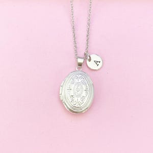May include: A silver locket necklace with an ornate design and a small silver charm with the letter 'A' hanging from it. The necklace is on a pink background.