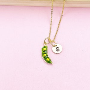 Peut inclure: Un collier dor&eacute; avec un charm en forme de gousse de pois et un petit charm circulaire avec la lettre "S". Le charm de la gousse de pois est vert avec trois pois visibles. Le collier est pr&eacute;sent&eacute; sur un fond rose.