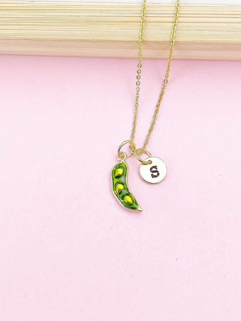 Peut inclure: Un collier dor&eacute; avec un charm en forme de gousse de pois et un petit charm circulaire avec la lettre "S". Le charm de la gousse de pois est vert avec trois pois visibles. Le collier est pr&eacute;sent&eacute; sur un fond rose.