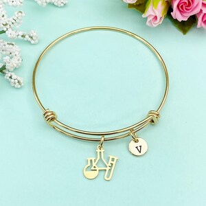 May include: Gold-colored bangle bracelet with two charms. One charm features a chemistry flask and test tube design, and the other is a small round charm with the letter "V". The bracelet is displayed on a light blue surface.
