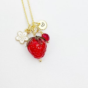May include: Gold chain necklace with a red glass strawberry charm, a white flower charm, and a gold charm with a heart engraved on it.