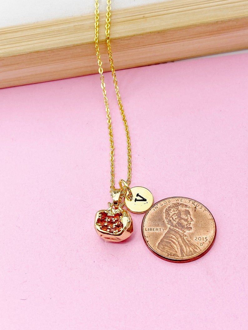 以下が含まれることがあります： 小さな金のザクロのチャームと金のイニシャルチャーム（「V」）が付いた金のチェーンネックレス。ネックレスは、アメリカの1セント硬貨の隣にあるピンクの表面に置かれています。