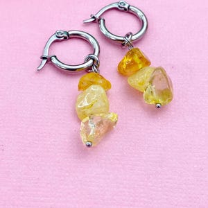 May include: A pair of hoop earrings with a silver finish. Each earring features a cluster of yellow and amber-colored gemstones. The earrings are displayed against a pink background, highlighting the jewelry's colors and design.