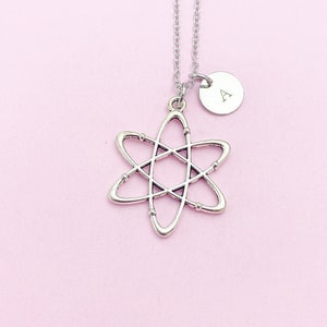 May include: A silver necklace with an atom pendant and a small round charm with the letter 'A' engraved on it. The necklace is on a pink background.