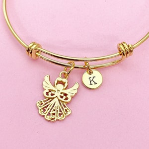 K&ouml;nnte beinhalten: Ein goldenes Armreif-Armband mit einem zarten Engelsanh&auml;nger und einem kleinen runden Anh&auml;nger mit dem eingravierten Buchstaben "K".
