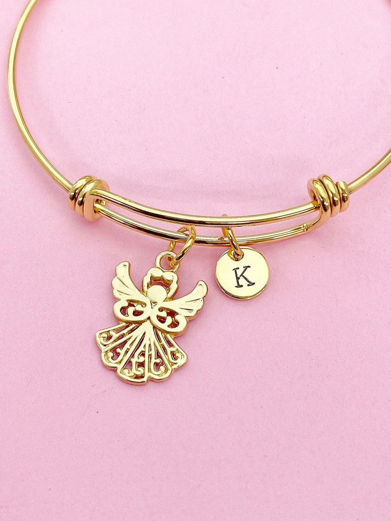 K&ouml;nnte beinhalten: Ein goldenes Armreif-Armband mit einem zarten Engelsanh&auml;nger und einem kleinen runden Anh&auml;nger mit dem eingravierten Buchstaben "K".
