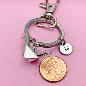 K&ouml;nnte beinhalten: Silberner Origami-Kranich-Schl&uuml;sselanh&auml;nger mit einem silbernen Anh&auml;nger mit dem Buchstaben M und einem US-Penny mit der Aufschrift "In God We Trust" und "Liberty".
