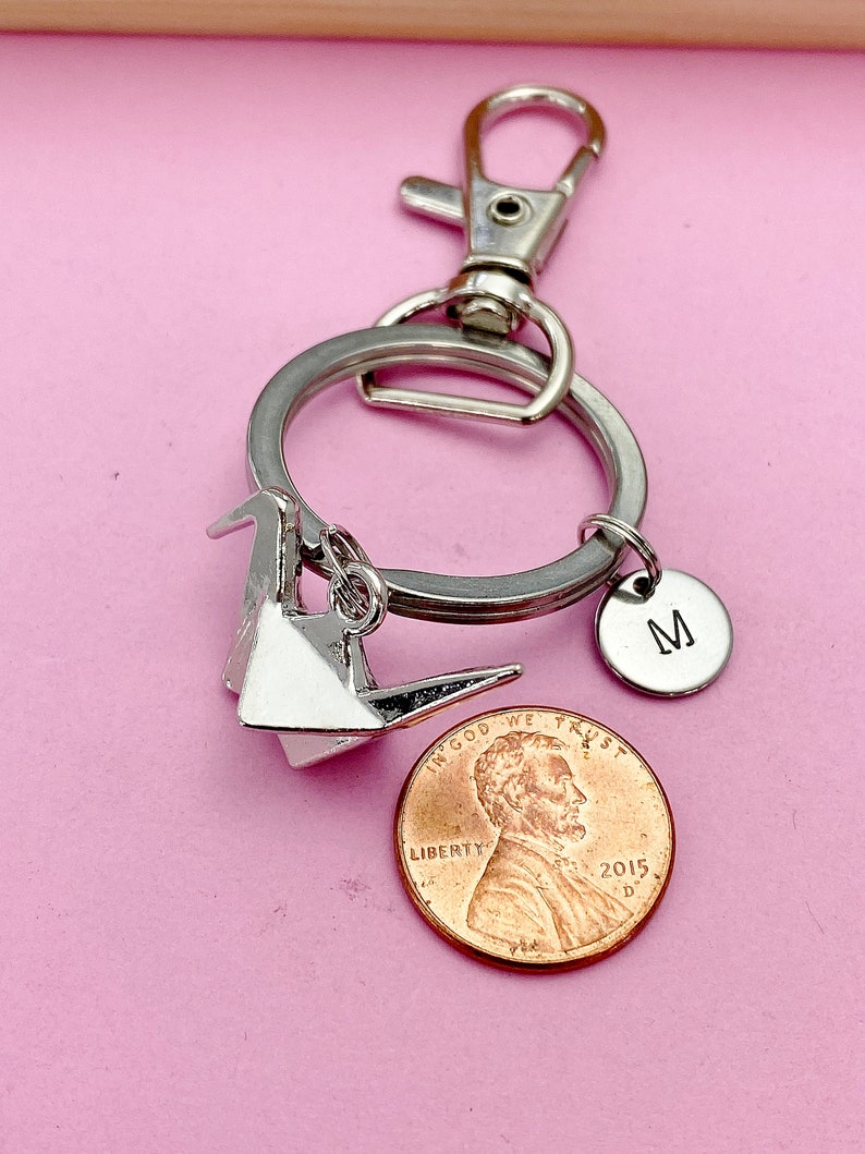 K&ouml;nnte beinhalten: Silberner Origami-Kranich-Schl&uuml;sselanh&auml;nger mit einem silbernen Anh&auml;nger mit dem Buchstaben M und einem US-Penny mit der Aufschrift "In God We Trust" und "Liberty".