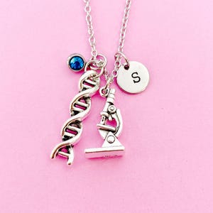 May include: Silver-toned necklace with three charms: a DNA helix, a microscope, and a circular charm with the letter "S". A blue gemstone is also attached. The necklace is displayed against a pink background.