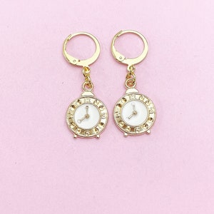 May include: A pair of gold-tone earrings with a white clock face design. The clock face has black Roman numeral markings and gold-tone hands.