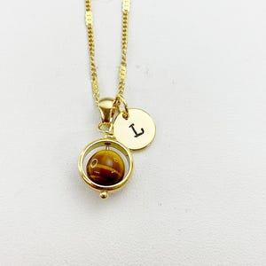 May include: Gold chain necklace with a spinning tiger's eye stone pendant and a small gold disc charm with the letter 'L' engraved on it.