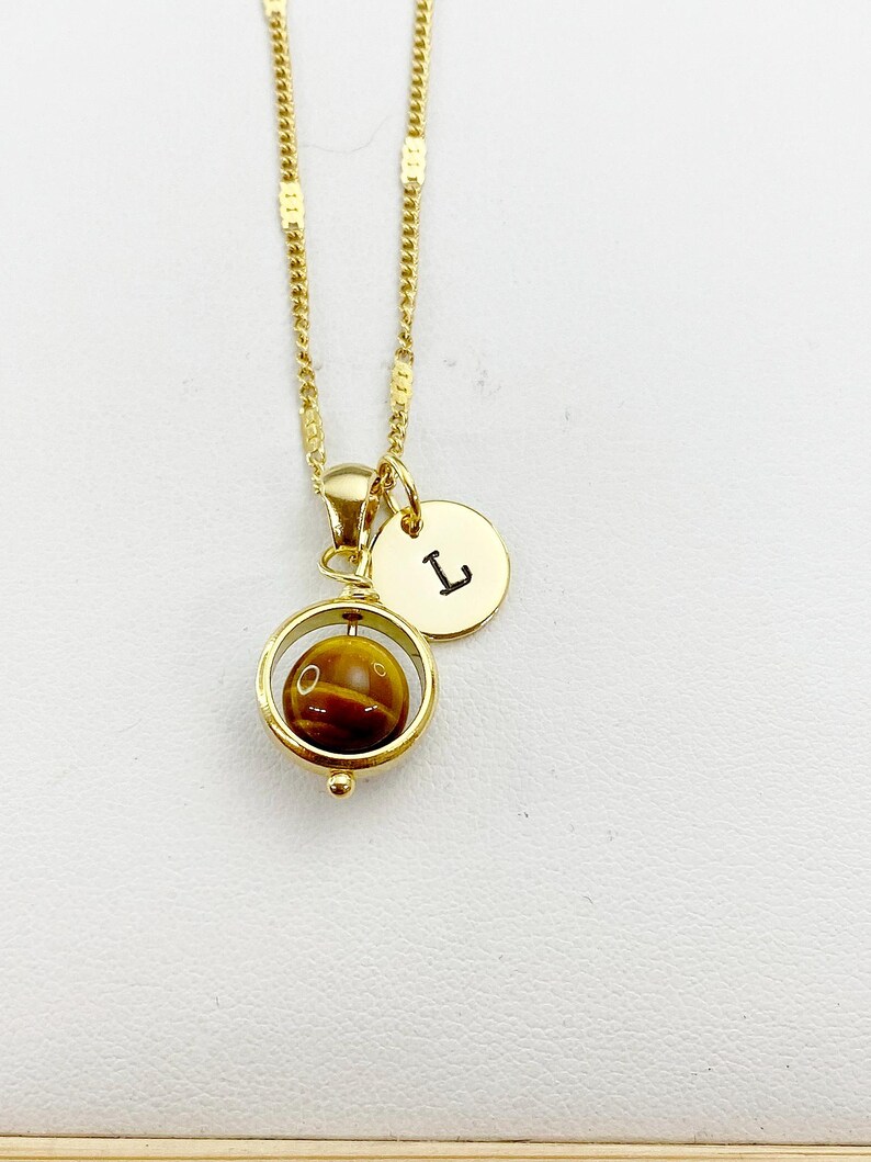 May include: Gold chain necklace with a spinning tiger's eye stone pendant and a small gold disc charm with the letter 'L' engraved on it.