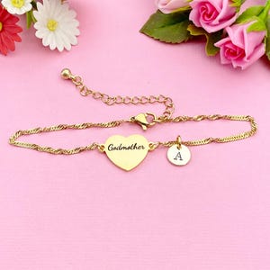 May include: Gold-toned bracelet with a heart-shaped charm engraved with "Godmother" and a circular charm with the letter "A". The bracelet has a delicate chain design and a lobster clasp. The background is pink with floral accents.