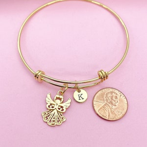 K&ouml;nnte beinhalten: Goldenes Armreif-Armband mit einem Anh&auml;nger eines Engels und einem Anh&auml;nger mit dem Buchstaben K. Das Armband hat auch einen Anh&auml;nger eines 1-Cent-St&uuml;cks aus dem Jahr 2015.