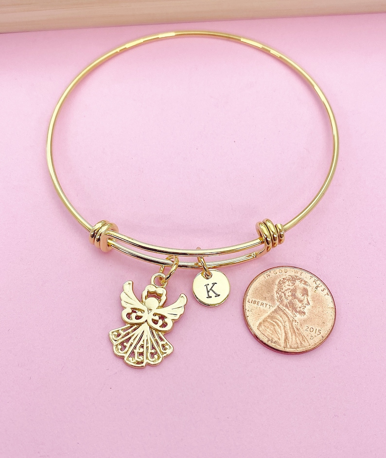 K&ouml;nnte beinhalten: Goldenes Armreif-Armband mit einem Anh&auml;nger eines Engels und einem Anh&auml;nger mit dem Buchstaben K. Das Armband hat auch einen Anh&auml;nger eines 1-Cent-St&uuml;cks aus dem Jahr 2015.