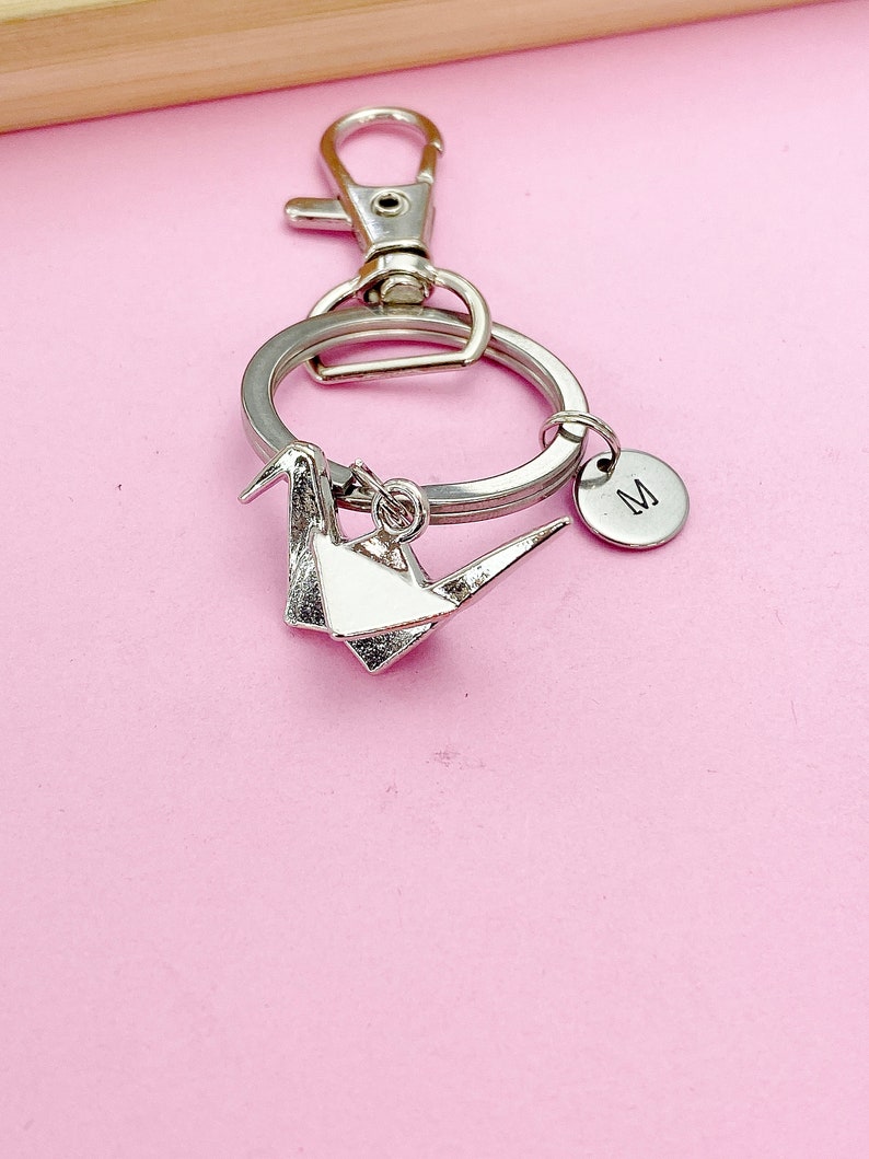 K&ouml;nnte beinhalten: Silberner Origami-Kranich-Schl&uuml;sselanh&auml;nger mit einem silbernen geteilten Ring und einem silbernen Karabinerhaken. Ein kleiner silberner Anh&auml;nger mit dem Buchstaben "M" ist am geteilten Ring befestigt.