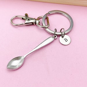 May include: Silver spoon keychain with a silver split ring and a silver charm with the letter "B" engraved on it. The keychain is on a pink background.