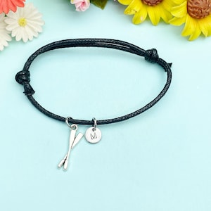 May include: Black adjustable bracelet with silver-tone charms. The charms include a pair of crossed oars and a round disc with the letter "M". The bracelet is displayed on a light blue surface, with colorful flowers in the background.
