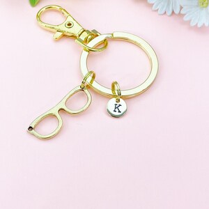 May include: Gold-colored keychain with a glasses charm and a round charm with the letter "K". The keychain has a large ring and a clasp. The background is a light pink with flowers.