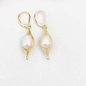 May include: A pair of gold-colored wire-wrapped earrings, each featuring a large, white, oval-shaped pearl. The pearls are encased in a delicate, woven wire design, with a lever-back closure. The earrings are displayed against a white background.