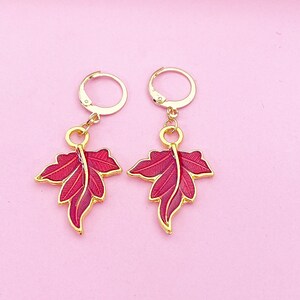 May include: A pair of gold hoop earrings with red enamel leaf charms. The leaves have a detailed design and are a vibrant red color.