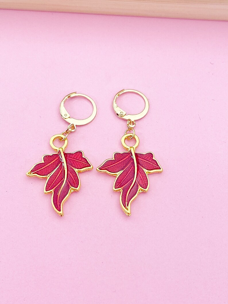 May include: A pair of gold hoop earrings with red enamel leaf charms. The leaves have a detailed design and are a vibrant red color.