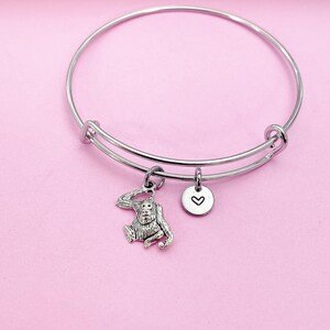 May include: A silver bangle bracelet with a charm of an orangutan and a small heart charm. The bracelet is on a pink background.
