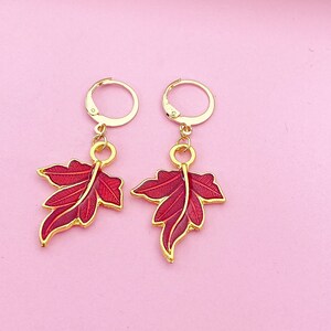 May include: A pair of gold hoop earrings with red leaf charms. The leaves have a detailed design and a glossy finish.