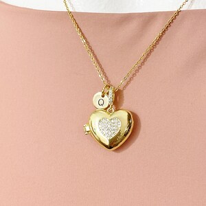 May include: Gold-colored heart-shaped locket necklace with a small round charm and a delicate chain. The heart locket is embellished with a sparkling crystal-like design in the center. The round charm has the letter "Q" on it.
