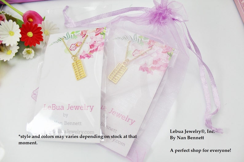Peut inclure: Deux colliers abacus dor&eacute;s sur des cartes blanches, emball&eacute;s dans des sacs en plastique transparent et pr&eacute;sent&eacute;s dans un sac cadeau lavande transparent. Les cartes pr&eacute;sentent le texte "LeBua Jewelry" et des motifs floraux. L'image comprend &eacute;galement le texte "LeBua Jewelry&reg;, Inc. By Nan Bennett".