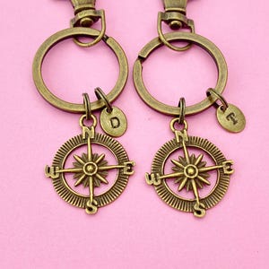 May include: Two gold-toned compass keychains with a small charm with the letters "D" and "T" on each. The keychains are on a pink background.