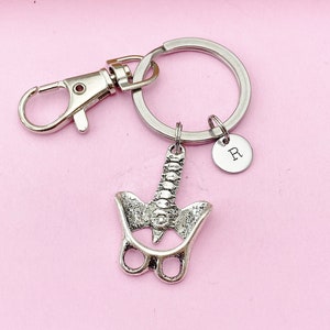 May include: Silver keychain featuring a detailed anatomical model of a human vertebra, attached to a silver split ring with a silver lobster clasp and a small silver charm with the letter 'R' engraved on it.