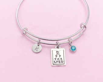 May include: Silver-toned bangle bracelet with three charms. One charm is a small disc with the letter "K", another is a miniature eye chart, and the third is a small, round, blue gemstone. The bracelet is on a pink background.