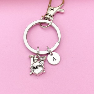 May include: Silver keychain with a drum charm and a silver charm with the letter A. The keychain is on a pink background.