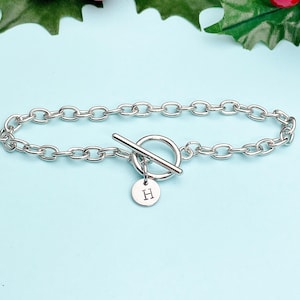 May include: A silver-toned chain link bracelet featuring a toggle clasp closure. The bracelet includes a small, round, flat charm dangling from the circular ring of the toggle, which is engraved with the capital letter H in a simple font.
