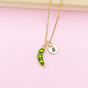 Peut inclure: Un collier dor&eacute; avec un pendentif en forme de gousse de pois et un charme circulaire avec la lettre "S". La gousse de pois est verte avec trois petits pois jaunes &agrave; l'int&eacute;rieur. Le collier est sur fond rose.