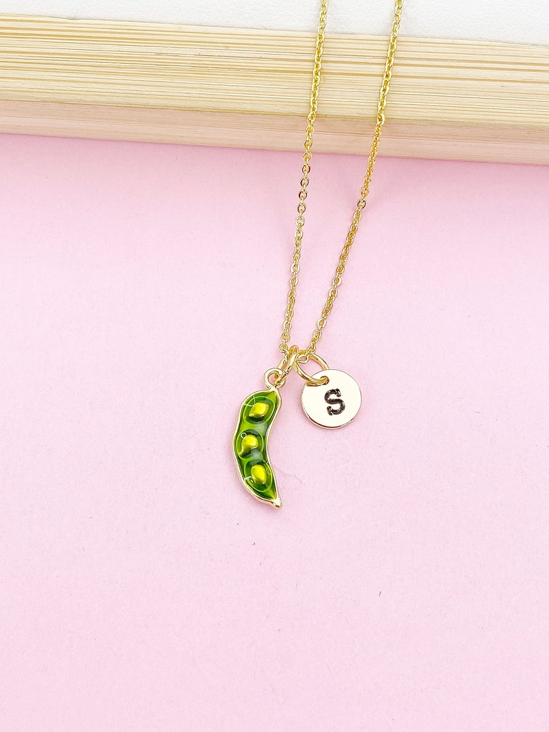 Peut inclure: Un collier dor&eacute; avec un pendentif en forme de gousse de pois et un charme circulaire avec la lettre "S". La gousse de pois est verte avec trois petits pois jaunes &agrave; l'int&eacute;rieur. Le collier est sur fond rose.