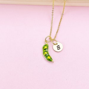 Peut inclure: Un collier dor&eacute; avec un pendentif en forme de gousse de pois verte et un petit m&eacute;daillon rond avec la lettre "S". La gousse de pois est verte avec des d&eacute;tails jaunes. Le collier est sur fond rose.