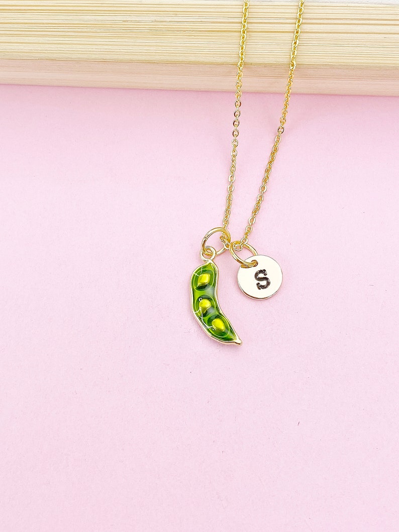Peut inclure: Un collier dor&eacute; avec un pendentif en forme de gousse de pois verte et un petit m&eacute;daillon rond avec la lettre "S". La gousse de pois est verte avec des d&eacute;tails jaunes. Le collier est sur fond rose.