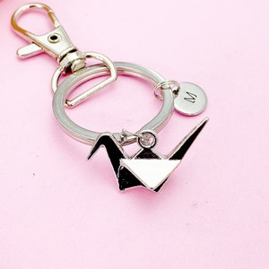 K&ouml;nnte beinhalten: Silberner Origami-Kranich-Schl&uuml;sselanh&auml;nger mit einem silbernen Schl&uuml;sselring und einem silbernen Anh&auml;nger mit dem eingravierten Buchstaben "M".