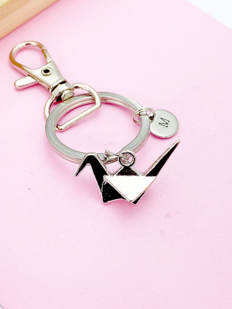 K&ouml;nnte beinhalten: Silberner Origami-Kranich-Schl&uuml;sselanh&auml;nger mit einem silbernen Schl&uuml;sselring und einem silbernen Anh&auml;nger mit dem eingravierten Buchstaben "M".