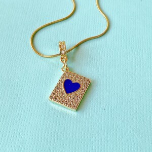 May include: A gold-tone necklace featuring a rectangular pendant encrusted with small clear crystals and a central blue heart-shaped enamel inlay. The pendant hangs from a delicate gold-tone snake chain, with a small crystal-set bail at the top.